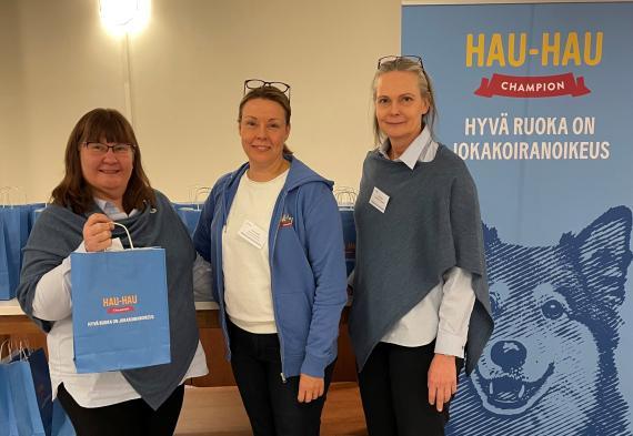 Kolme naista Hau-Hau Championin roll-upin edessä jakamassa Hau-Hau -kasseja