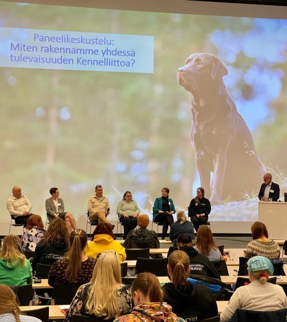 Näkymä täyden luentosalin eteen lavalle, jossa istuu rivissä tuoleilla kuusi ihmistä. Taustalla koira-aiheinen kuva ja teksti paneelikeskustelusta.