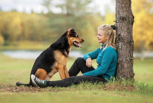 Koira istuu vastapäätä puuhun nojaavaa hymyilevää naista ja katsovat toisiaan silmiin