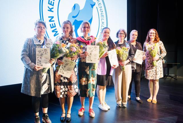 Parhaan jalostuksen tavoiteohjelman voittajat rivissä kukkineen ja diplomeineen, taustalla Kennelliiton logo seinälle heijastettuna.