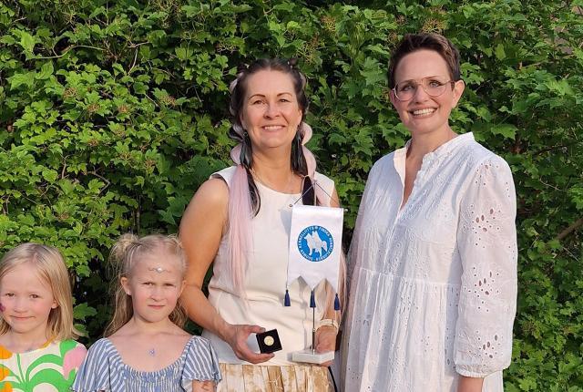 Sanna Törnroos Kennelliiton viirin ja Pro Iuvenes -ansiomerkin kanssa vehreän pensaan edessä, vieressä Molli Nyman ja kaksi pientä tyttöä.