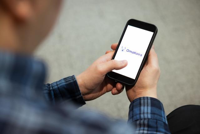 Henkilön kädessä oleva puhelin, jonka näytöllä näkyy Kennelliiton Omakoira-palvelun logo. Logossa on piirretty iloinen suomenpystykorvan naama.