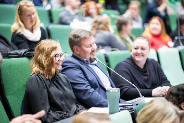 Kennelliiton valtuuston jäseniä istumassa valtuuston kokouksessa, kuvassa näkyy kaksi naista ja mies tuoleissaan hymyillen juttelemassa keskenään, taustalla näkyy muita valtuuston jäseniä.