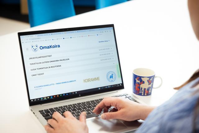 Tietokoneen näytöllä näkyy Omakoira-palvelun aloitussivu, sinipaitainen henkilö istuu koneella ja hänestä näkyy vain osa olkapäästä ja näppäimistöllä olevat kädet