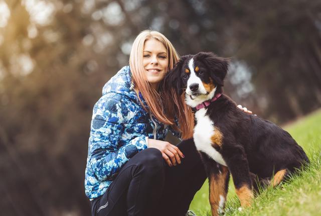 Nuori tyttö ja berninpaimenkoiranpentu nurmikolla, tytöllä on vaaleanpunertavaksi värjätyt hiukset ja hän katsoo hymyillen kameraan.