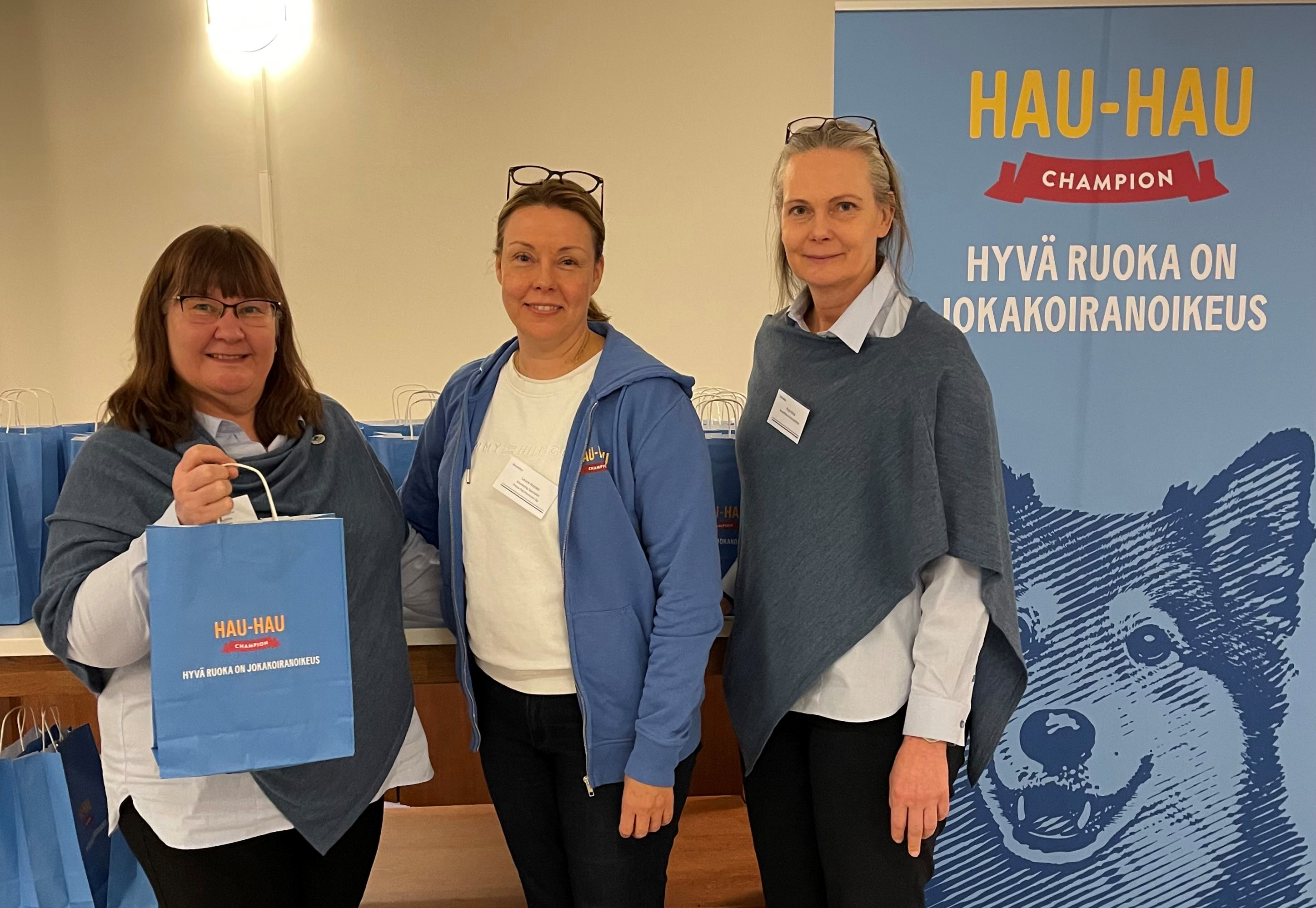 Kolme naista Hau-Hau Championin roll-upin edessä jakamassa Hau-Hau -kasseja